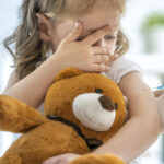 Angst vor Spritzen: Kleines Mädchen mit Teddybär im Arm schaut skeptisch auf die Spritze, die ihr der Arzt gerade verabreicht.